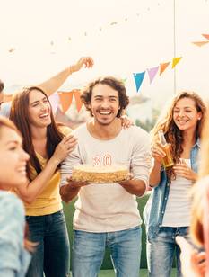Un groupe d’amis fête un anniversaire en plein air ; un homme souriant tient un gâteau orné du chiffre 30, entouré de décorations colorées et de convives joyeux.