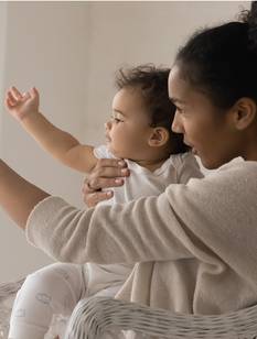 Une mère montre quelque chose à son bébé assis sur ses genoux dans un fauteuil en osier, dans une pièce lumineuse.