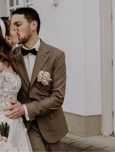 La mariée et le marié s'embrassent. En arrière-plan, on aperçoit le lieu de mariage. La mariée porte une robe blanche en dentelle et le marié un costume marron.