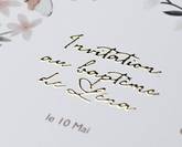 Gros plan sur un faire-part de baptême personnalisé, décoré de fleurs et lettré en doré, avec la date « le 10 mai ».