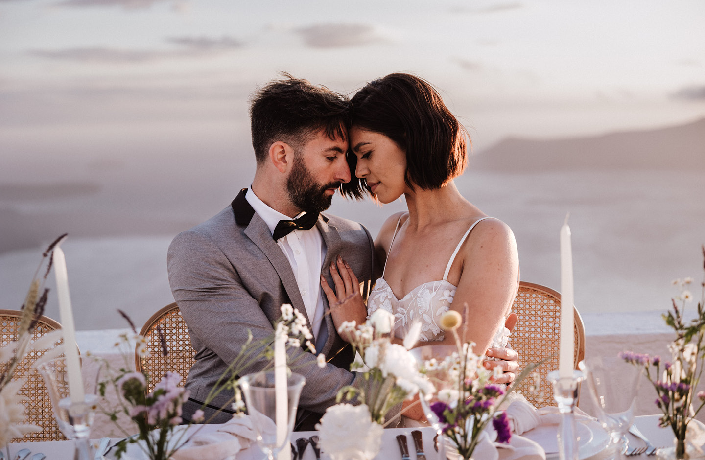 Couple de mariés se regardant tendrement à leur table de mariage, avec en arrière-plan flouté la mer Égée.