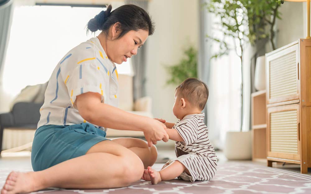 Une maman tient son bébé par les mains et l’aide à s’asseoir.