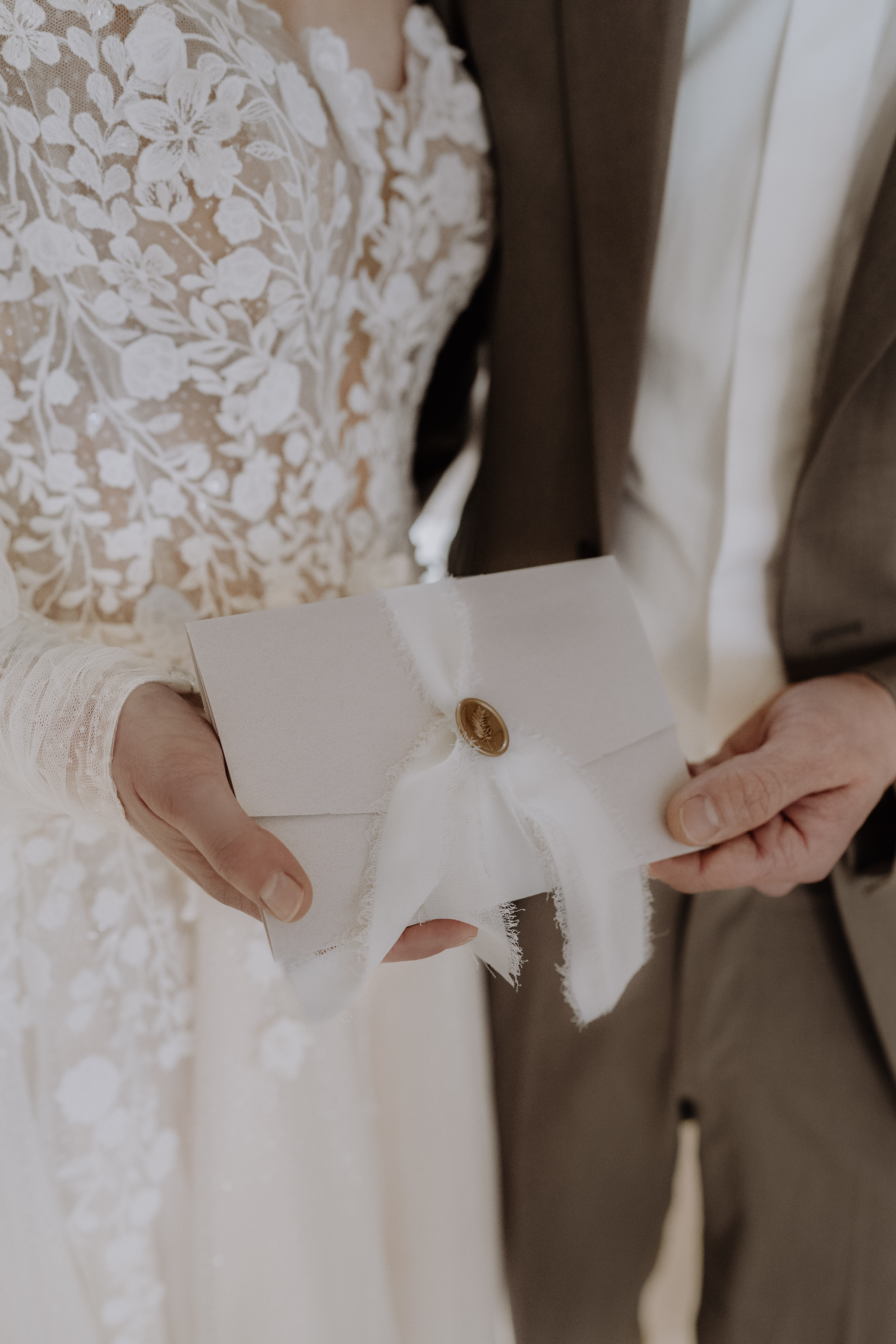 Détail visuel : la mariée et le marié tenant une invitation ornée d’un ruban et d’un sceau de cire