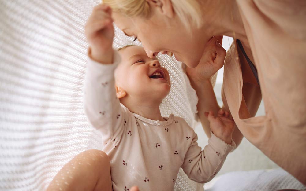 Une maman et son bébé sont assis par terre, riant ensemble sous une couverture.