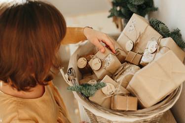 Christmas Advent calendar for kids. Girl wearing mustard linen dress looking at basket with advent gifts boxes on wooden cabinet. Eco friendly christmas