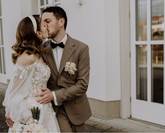 La mariée et le marié s'embrassent. En arrière-plan, on aperçoit le lieu de mariage. La mariée porte une robe blanche en dentelle et le marié un costume marron.