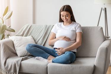 Beautiful young pregnant woman hugging her tummy while relaxing on couch at home, millennial expectant lady resting on sofa in living room, enjoying her pregnancy and future maternity, free space