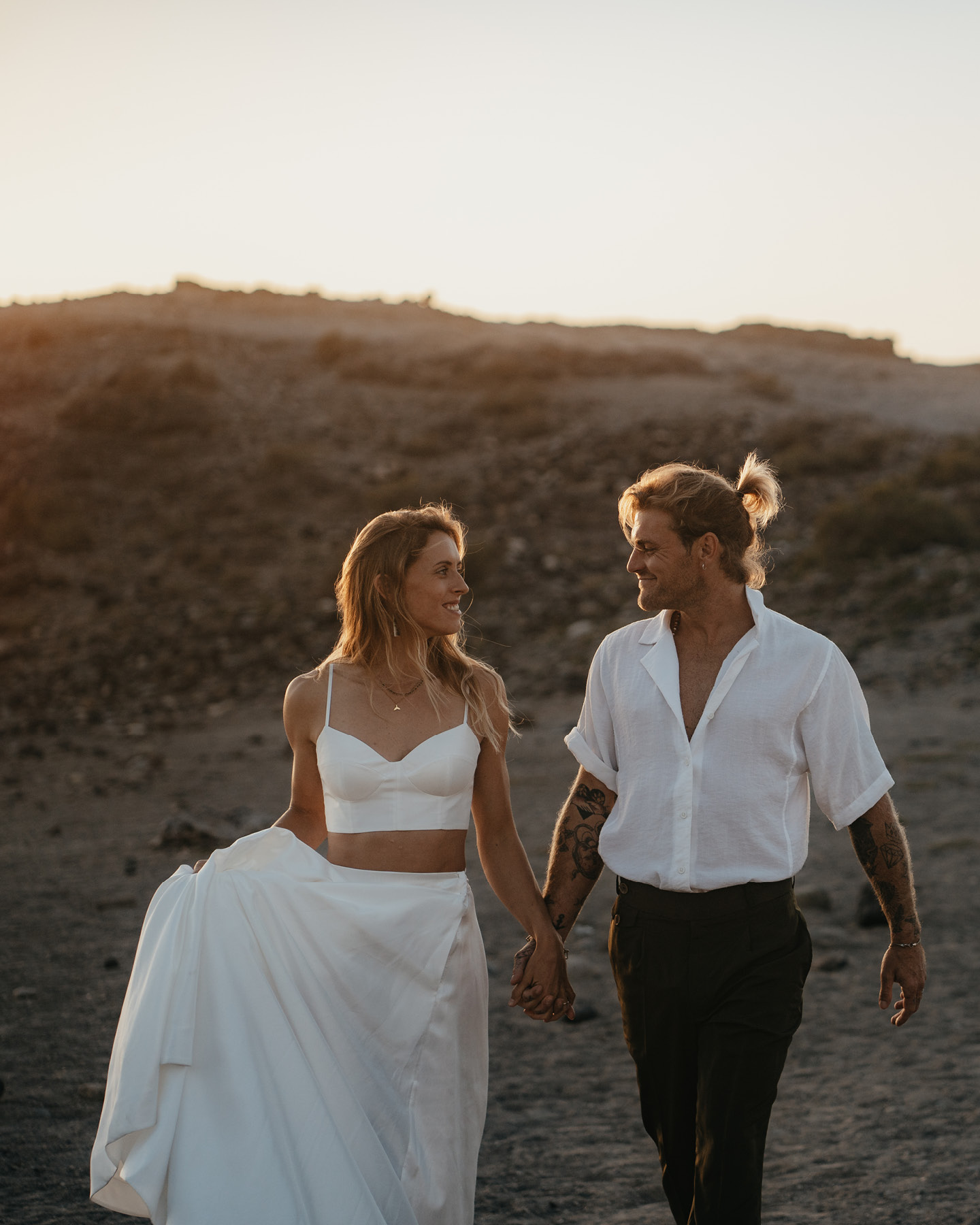 Couple de mariés, main dans la main sur la plage, vêtus d’une tenue à la fois élégante et décontractée